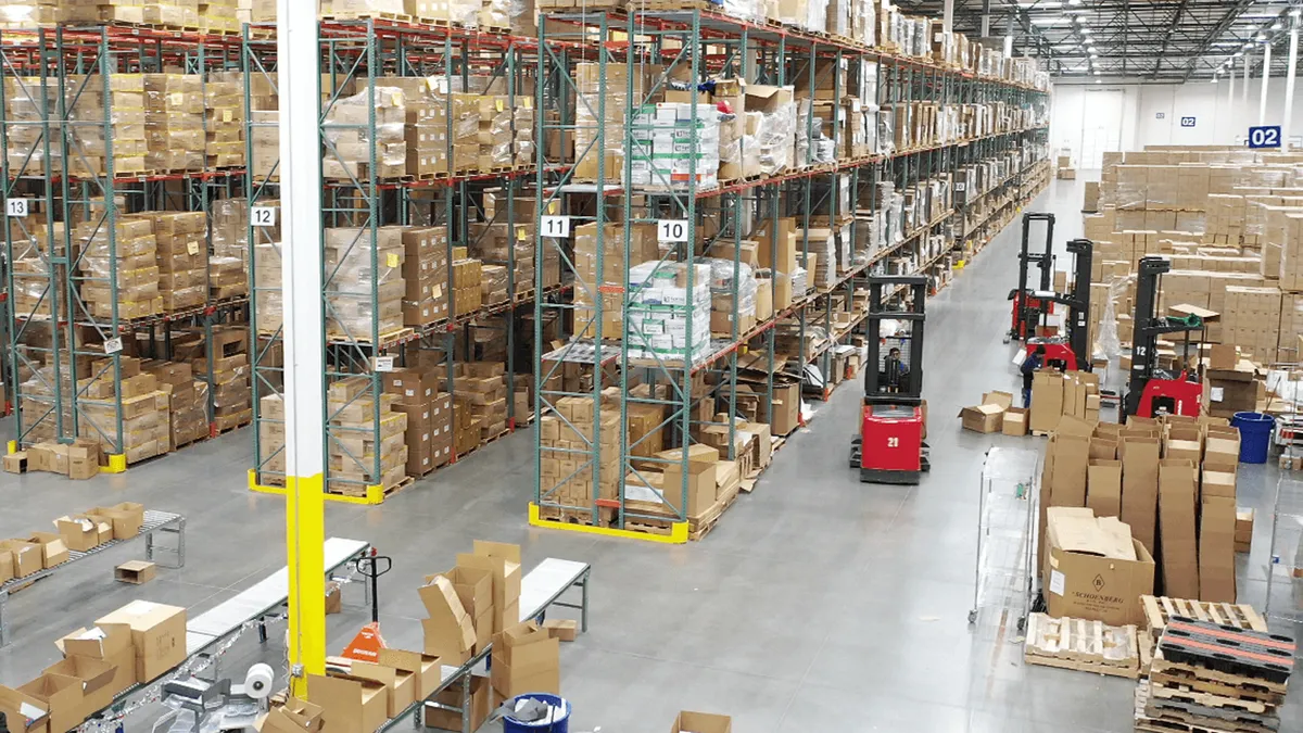 A person drives a forklift near rows of boxes that are stored on shelves in the interior of a warehouse.