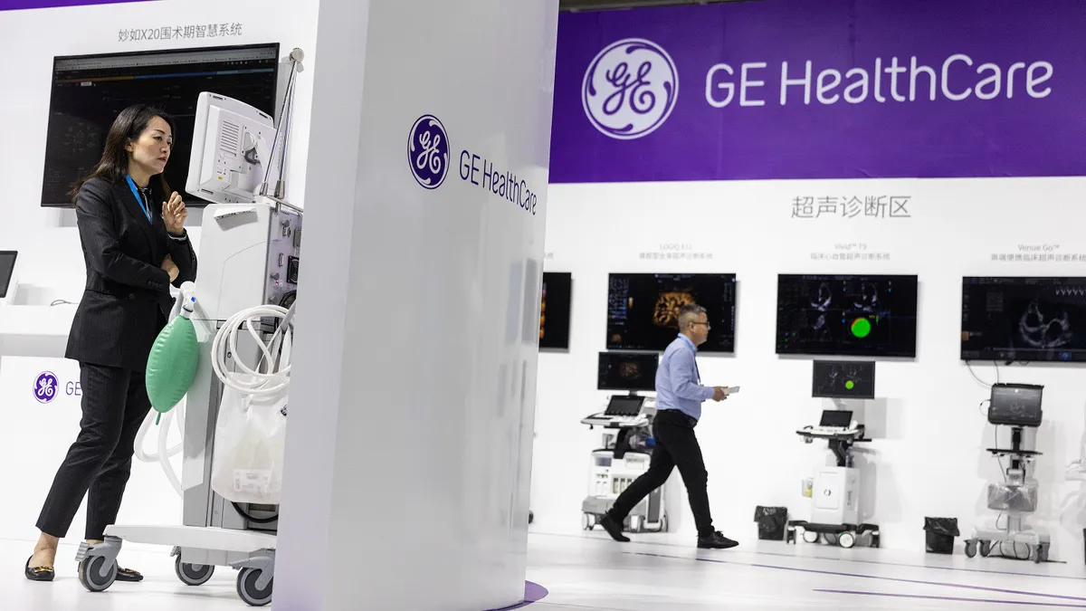 A person stands in front of the screen of a machine on wheels, near a sign that says "GE HealthCare"