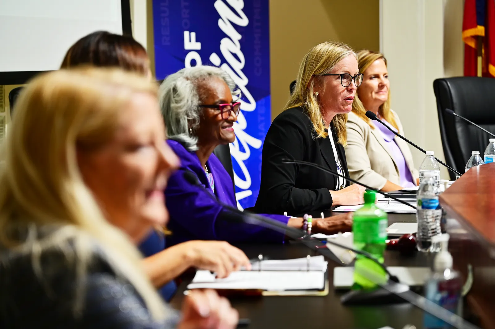 A multiethnic group of school board members are shown seated before microphones at a table during a meeting.