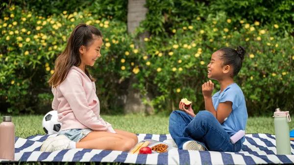 Two children sitting on a blanket in a yard, eating snacks and having a picnic.
