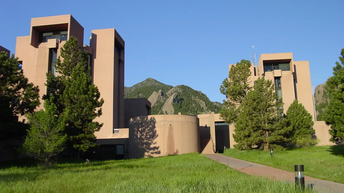 A building with two tall towers and a round center sits on a grassy site with mountains behind it.
