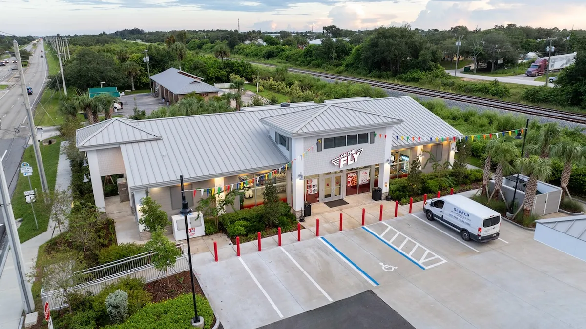 A photo of the exterior of a convenience store in Florida. The walls and roof are white, there are pennants from the building to two poles in the parking lot and there is a sign over the front door that reads "On the Fly."