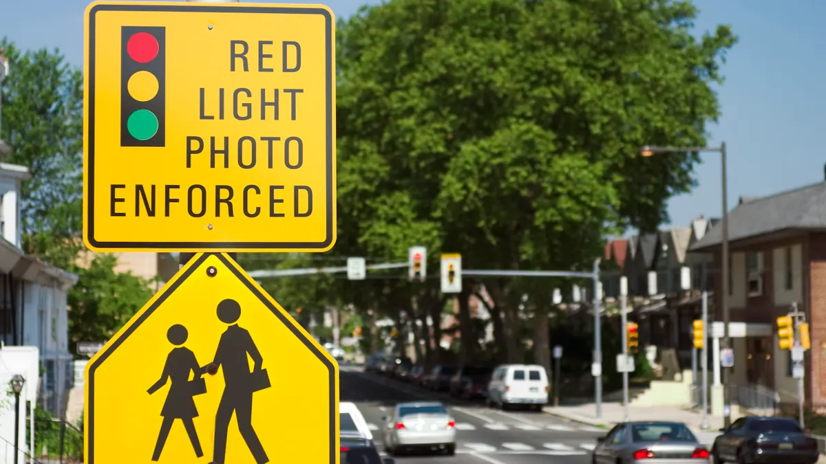 Yellow sign with graphic of a traffic signal reads "Red light photo enforced" over another yellow sign depicting pedestrians against a blurred backround of a city street.