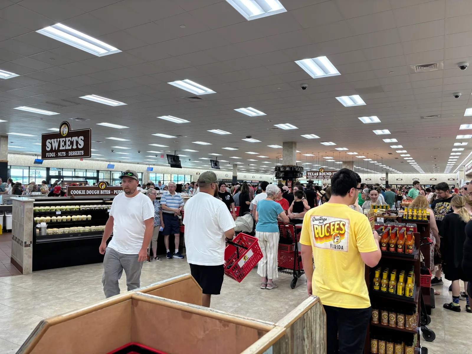 A photo of the interior of a large store. There is a crowd of people shopping. A sign hanging from the ceiling on the left side of the photo says "Sweets. Fudge. Nuts. Desserts."