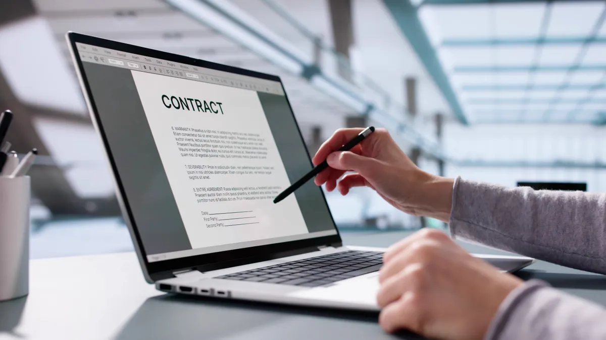A person's hand in front of a contract pictured on a laptop screen.
