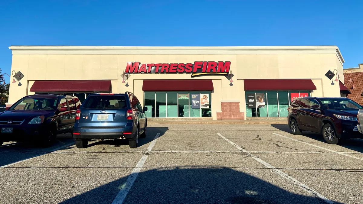 Cars are parked in front of a store with "Mattress Firm" in red capital letters, against a bright blue sky.