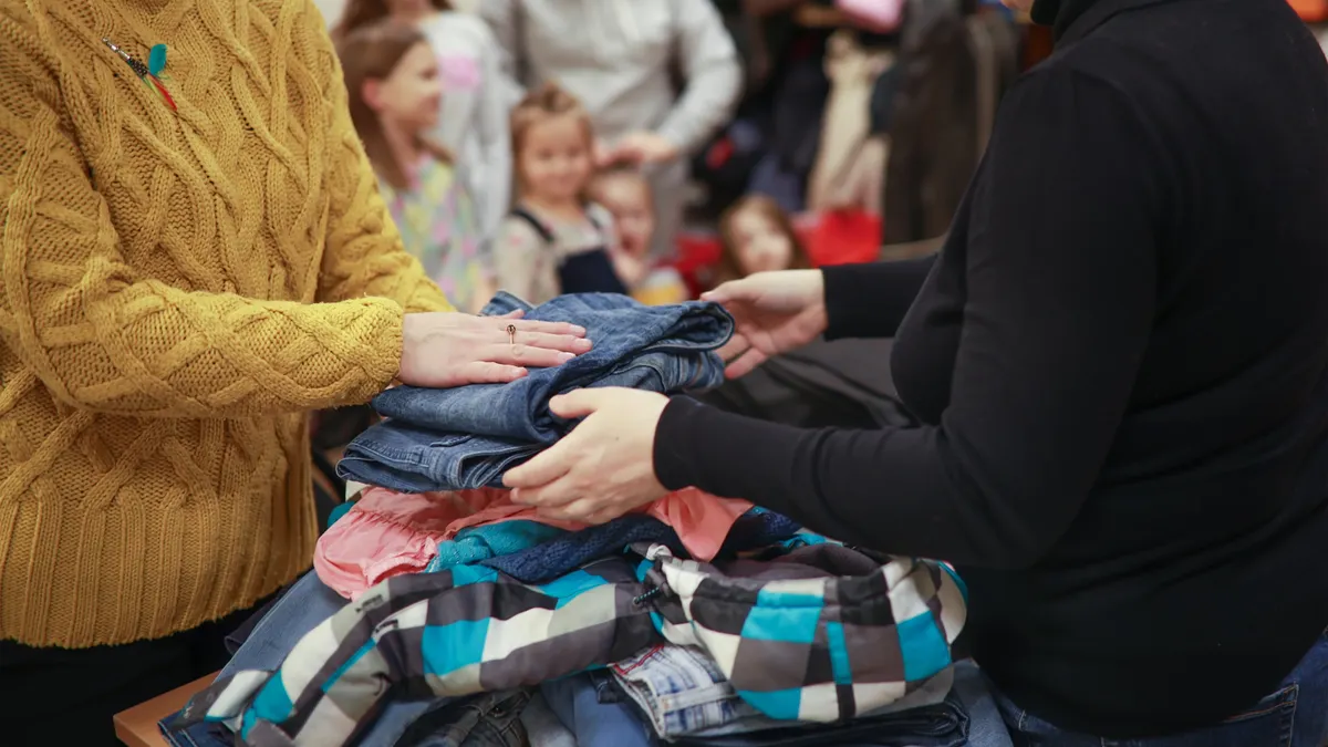 A woman volunteer helps another woman sort clothes on a table in a charity setting. Several children are in the background.