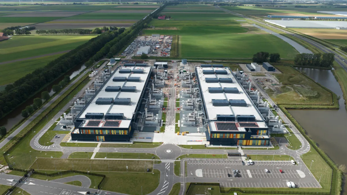 Aerial view of a large data center complex with two identical rectangular buildings surrounded by green fields and a clear sky, conveying modern industrial efficiency.
