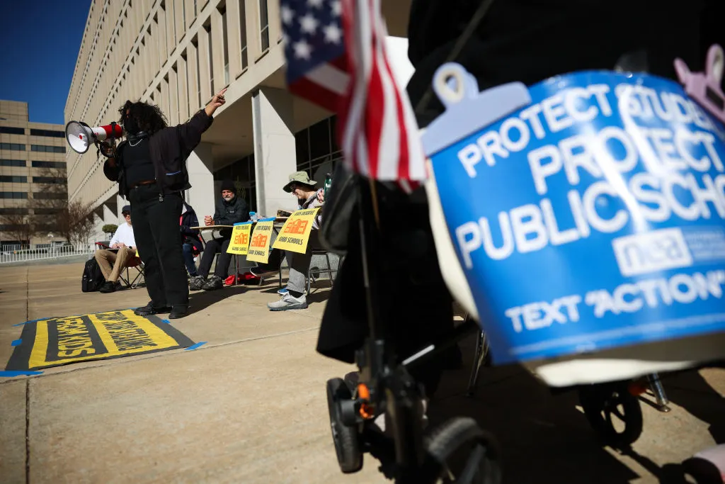 Several people are seated at desks outside of the U.S. Department of Education. Some people are standing and there are signs on the ground and being held. One sign says "Protect Students. Protect Public Schools."
