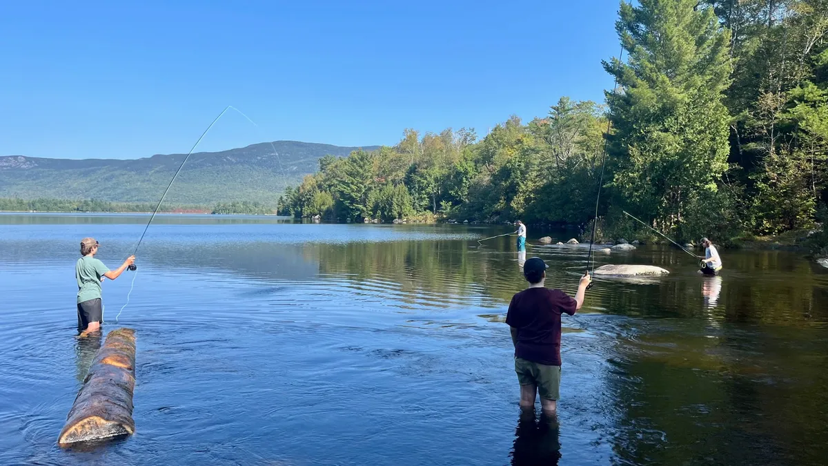 Several students stand knee-high in a waterway with fly-fishing rods and lines. Along the water are trees