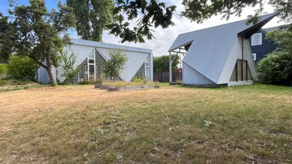 A field of grass on a corner lot with two small housing structures in the background.