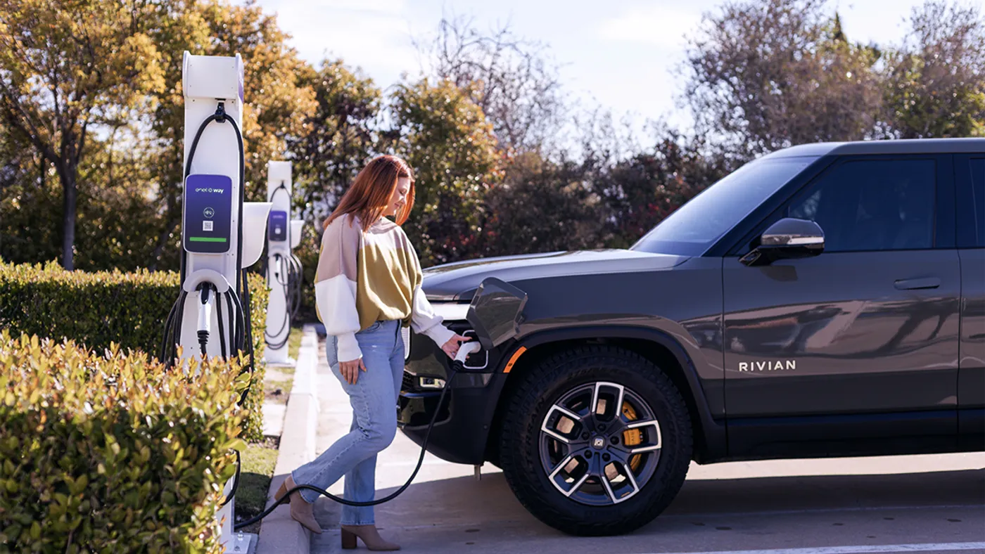 Several vehicles lined up along a curb are blurred while in the foreground a white and silver electric vehicle charger is in focus.