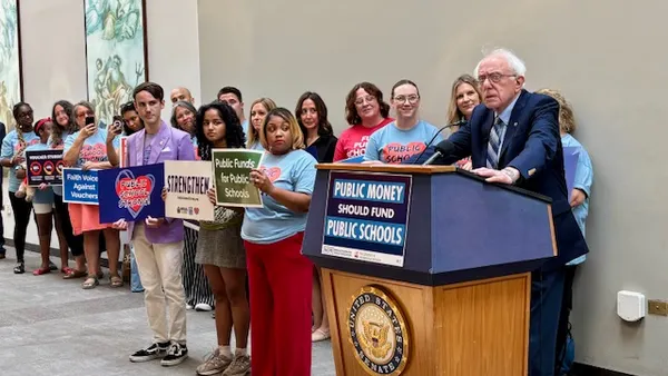 A person is standing at a podium with a microphone inside a large room. Behind the speaker are other people holding signs supporting public schools.
