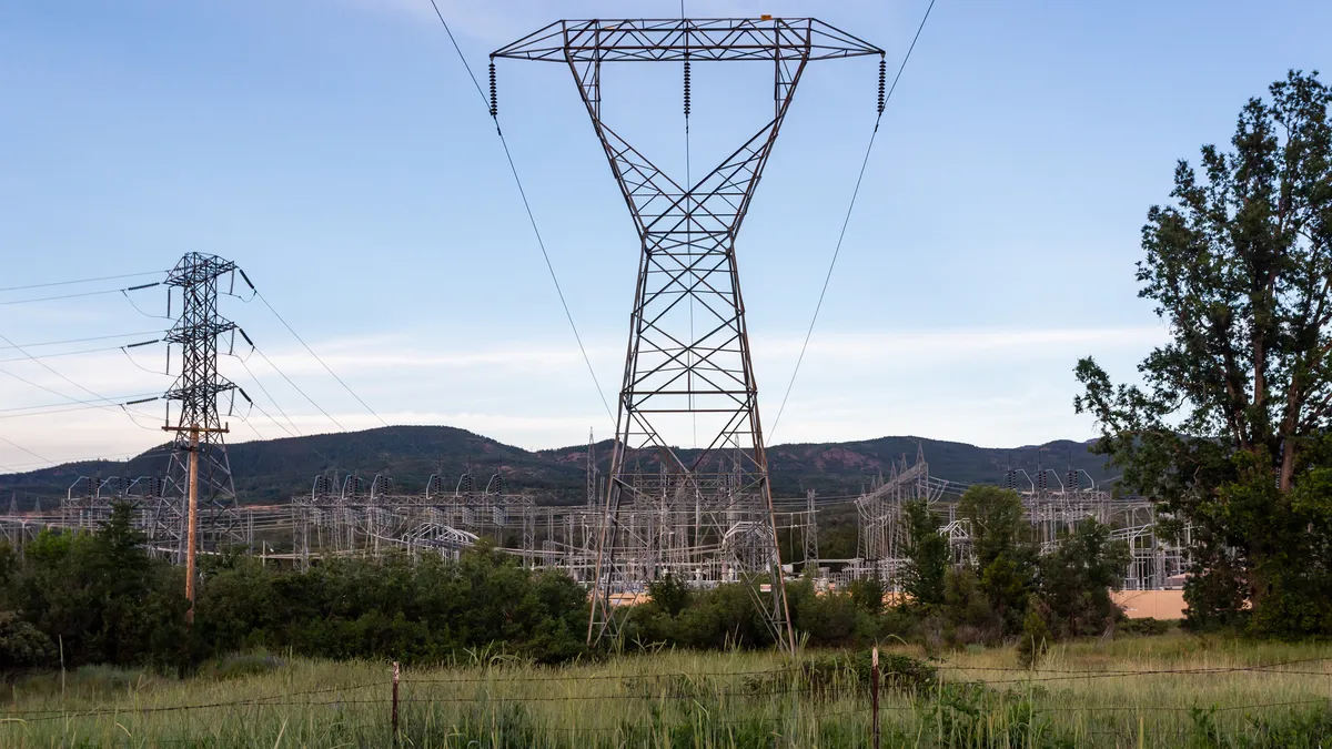 A transmission pylon stands in the middle of a field with electricity wires running to a substation behind it.