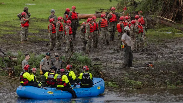 A crowd of rescue workers dressed in red vests and red helmets on the ground and workers dressed in yellow coats and red helmets in a blue boat on a river.