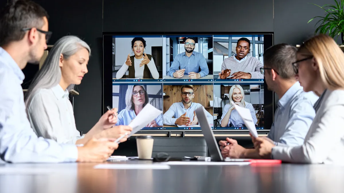 Online videoconference in meeting room with people sitting in modern office and colleagues on big screen monitor.