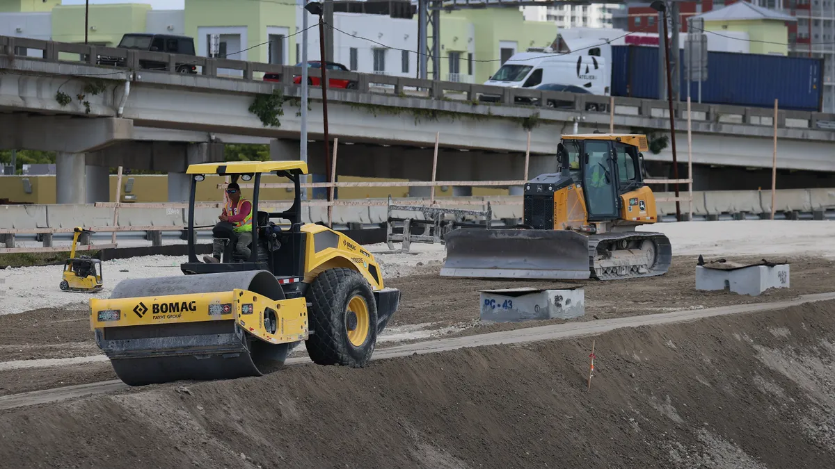 Construction worker in a yellow machine packs dirt on a jobsite with a bridge and more equipment in the background.