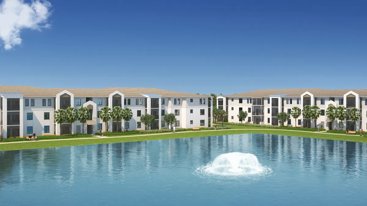 Tan, three-story apartment building with palm trees and a lake in the foreground.