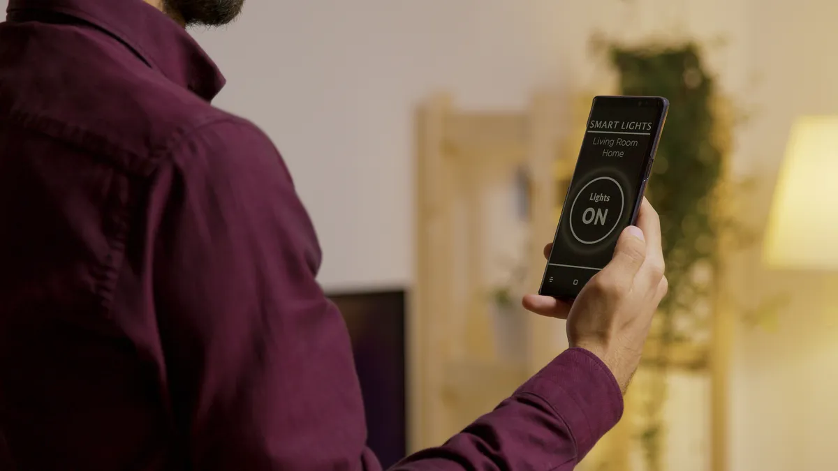 A person with a beard in a long-sleeved mauve dress shirt holds up a phone with a display that reads "Smart Lights - On." The person's face is not visible.