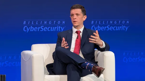 A man wearing a dark blue suit gestures with both hands while sitting in a plush white chair on a stage. Behind him, a backdrop displays a logo reading "Billington Cybersecurity."