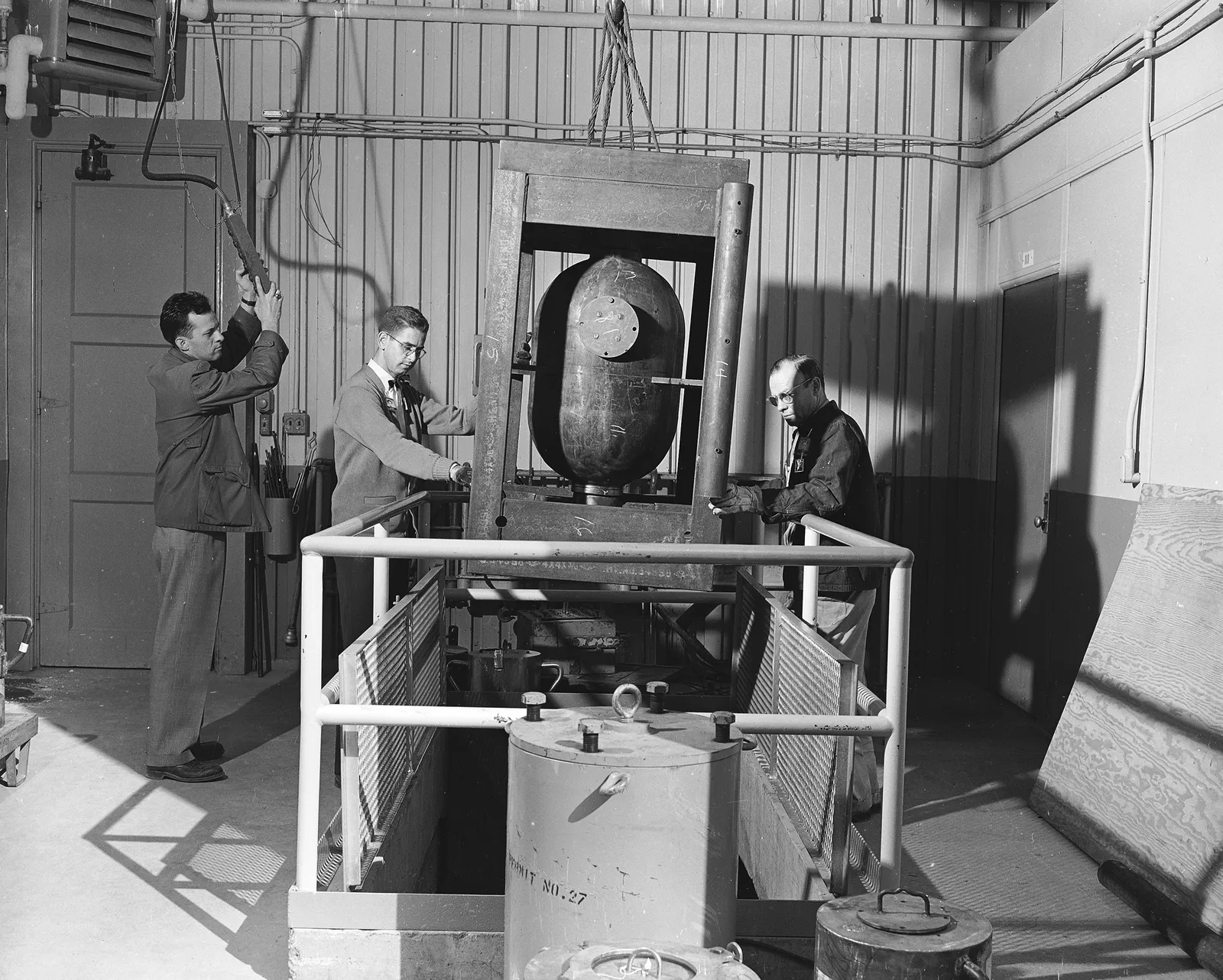 Three people stand around a suspended metal cylinder.