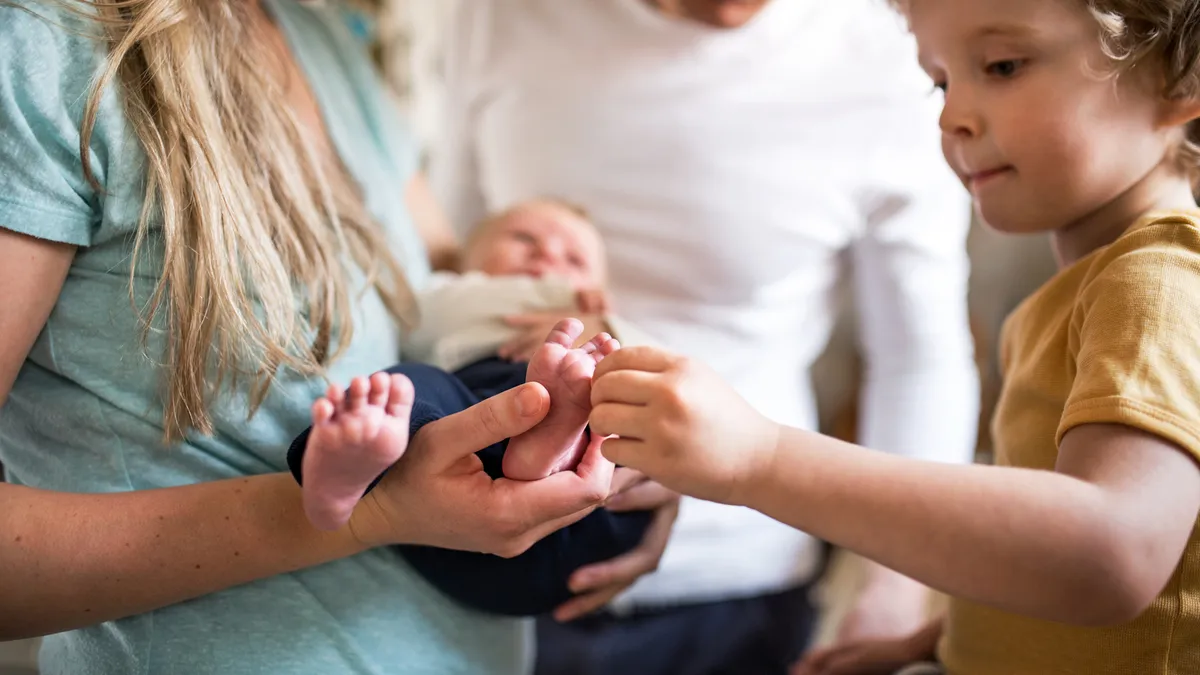 A midsection photo of two adults stand facing a toddler who is touching a baby on the foot in one of the adults arms.