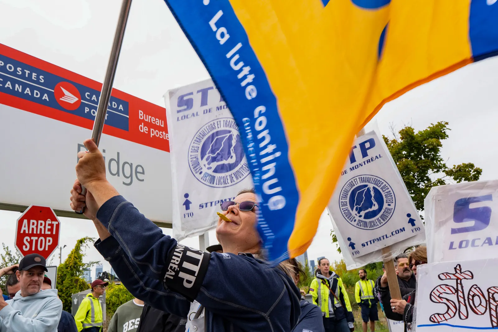 A group of people wave signs and flags reading “Arret,” “Stop,” and “La lutte continue.”