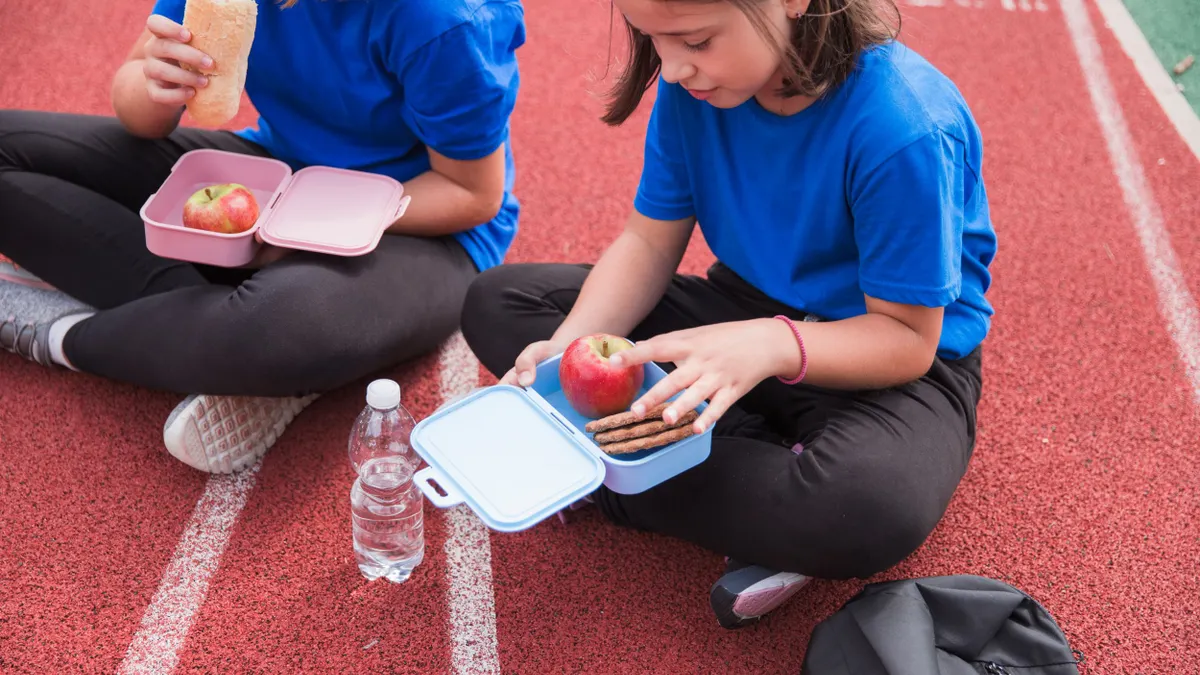 Two students sit together on a running track outside while eating from their lunch boxes.