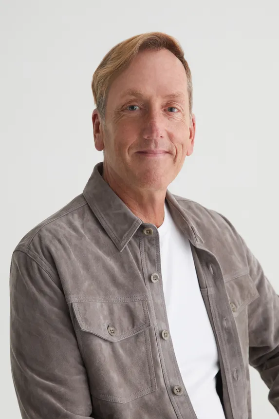 A man in a a white t-shirt, short blond hair, and a gray long sleeved shirt smiles for the camera.