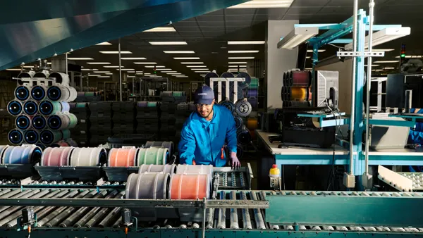 A person wearing a blue coverall suit, gloves, safety goggles and a blue Corning ball cap overseeing machinery.