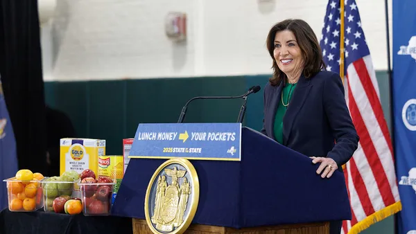 New York Gov. Kathy Hochul stands behind a podium next to a variety of food items as she announces a universal school meals proposal in a school cafeteria.