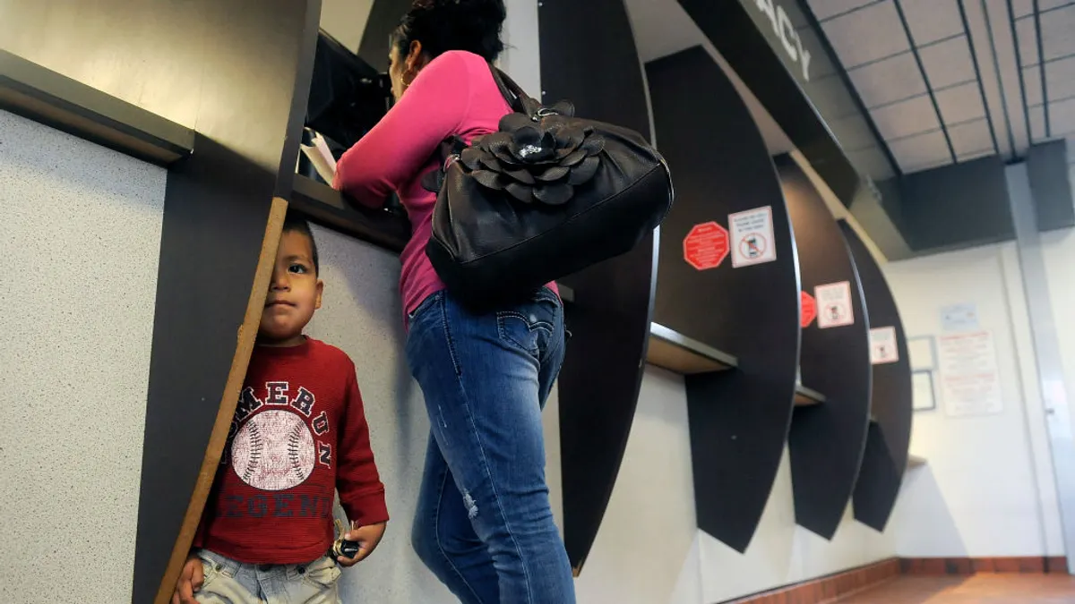 A woman in a pink shirt stands at a pharmacy counter while her son stands behind her, hiding behind a wall.