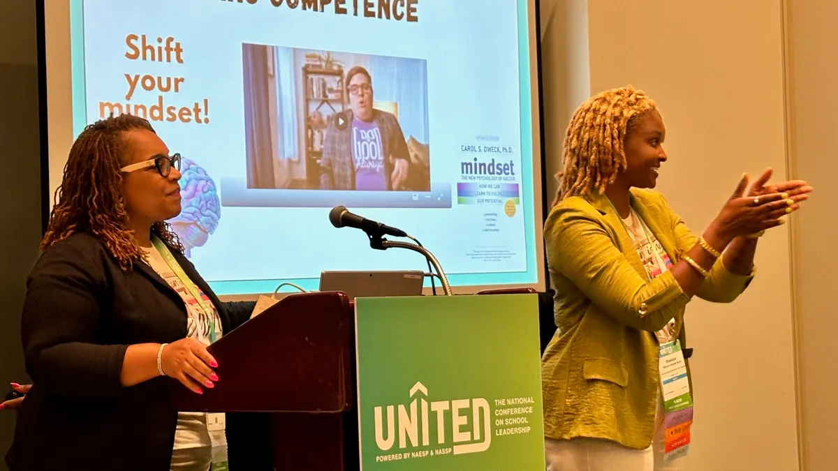 Two adults stand in a room. One is near a table with a podium and speaker. Another is standing in front of a screen that reads "presuming competence."