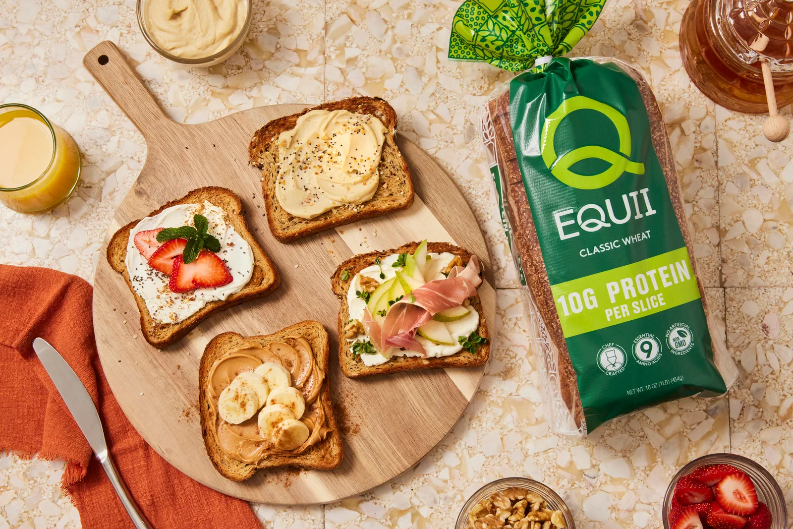 A round wooden cutting board with four slices of bread on it, each one with a spread and topping, sits on a peach colored stone counter with a bag of Equii bread to the right.