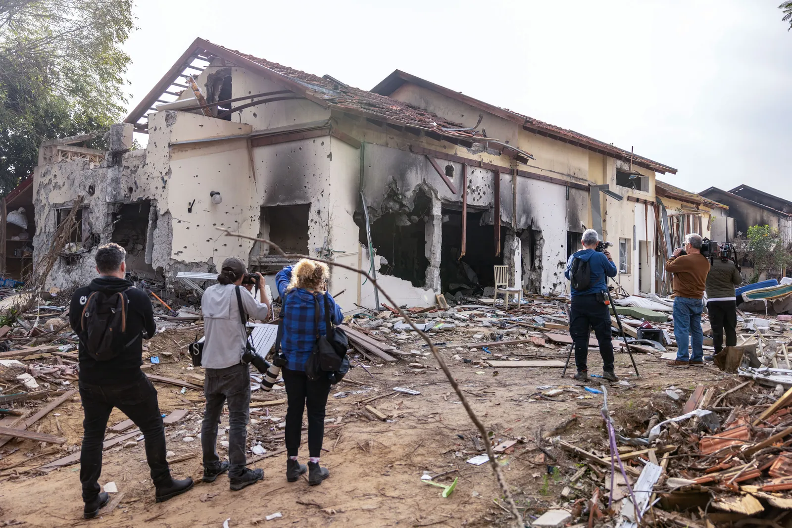 People stand with camera gear facing a home that is burned and destroyed, debris litter the ground.