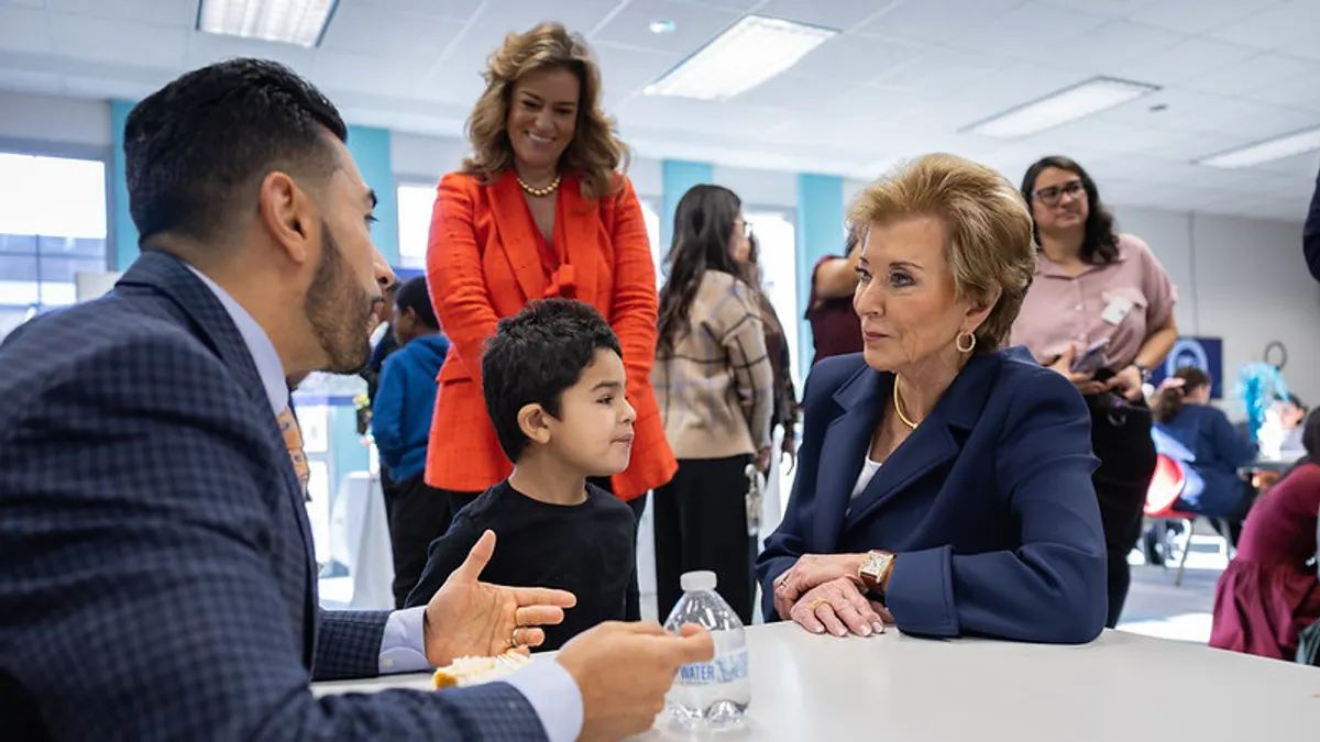 Two adults and a children are seated at a table in a school library. In the background another adult is standing and smiling and looking at the people around the table.