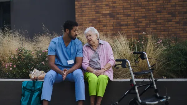 A healthcare worker in blue scrubs talks with an elderly woman sitting next to a walker outdoors.