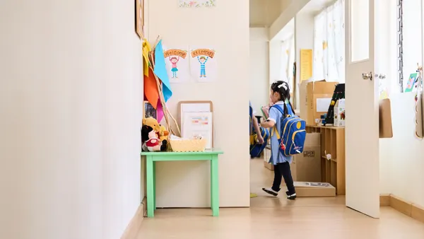A young student wearing a backpack and holding books enters a classroom. The student is shown from behind.