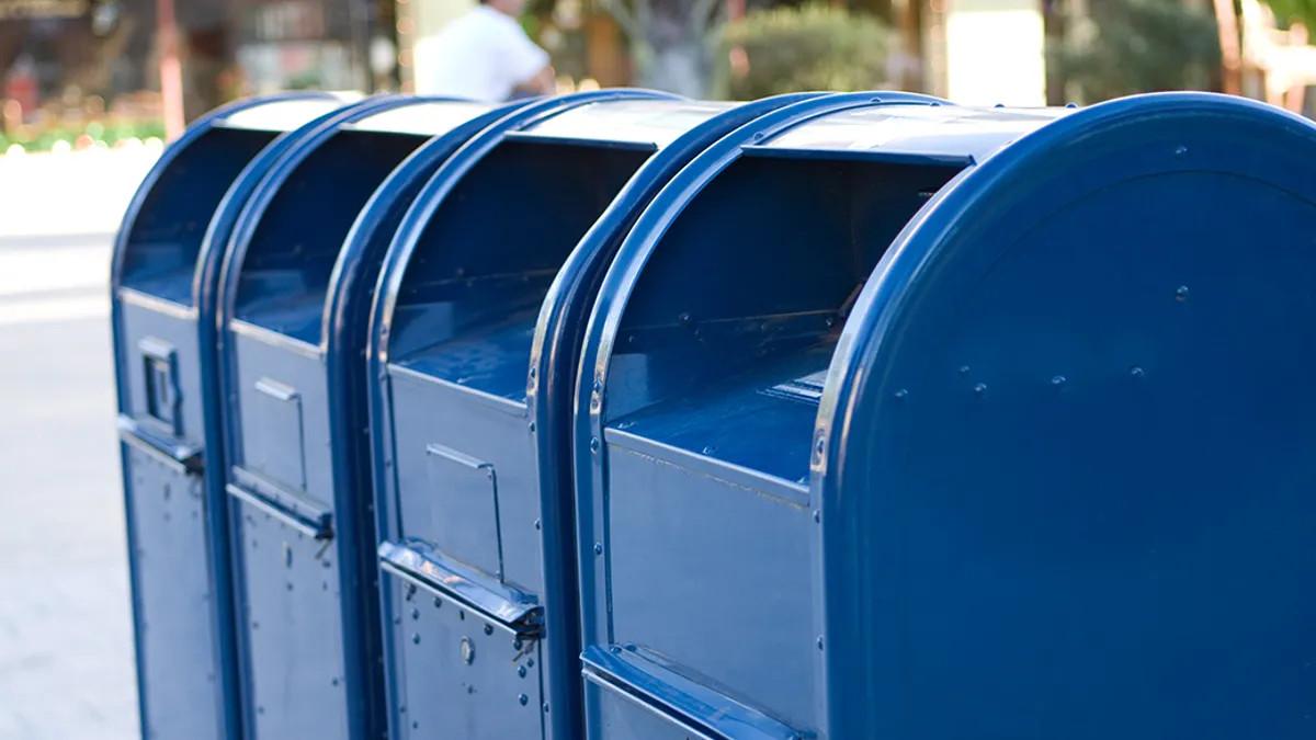 A row of four blue USPS mailboxes with open mail slots, set in an urban outdoor environment.