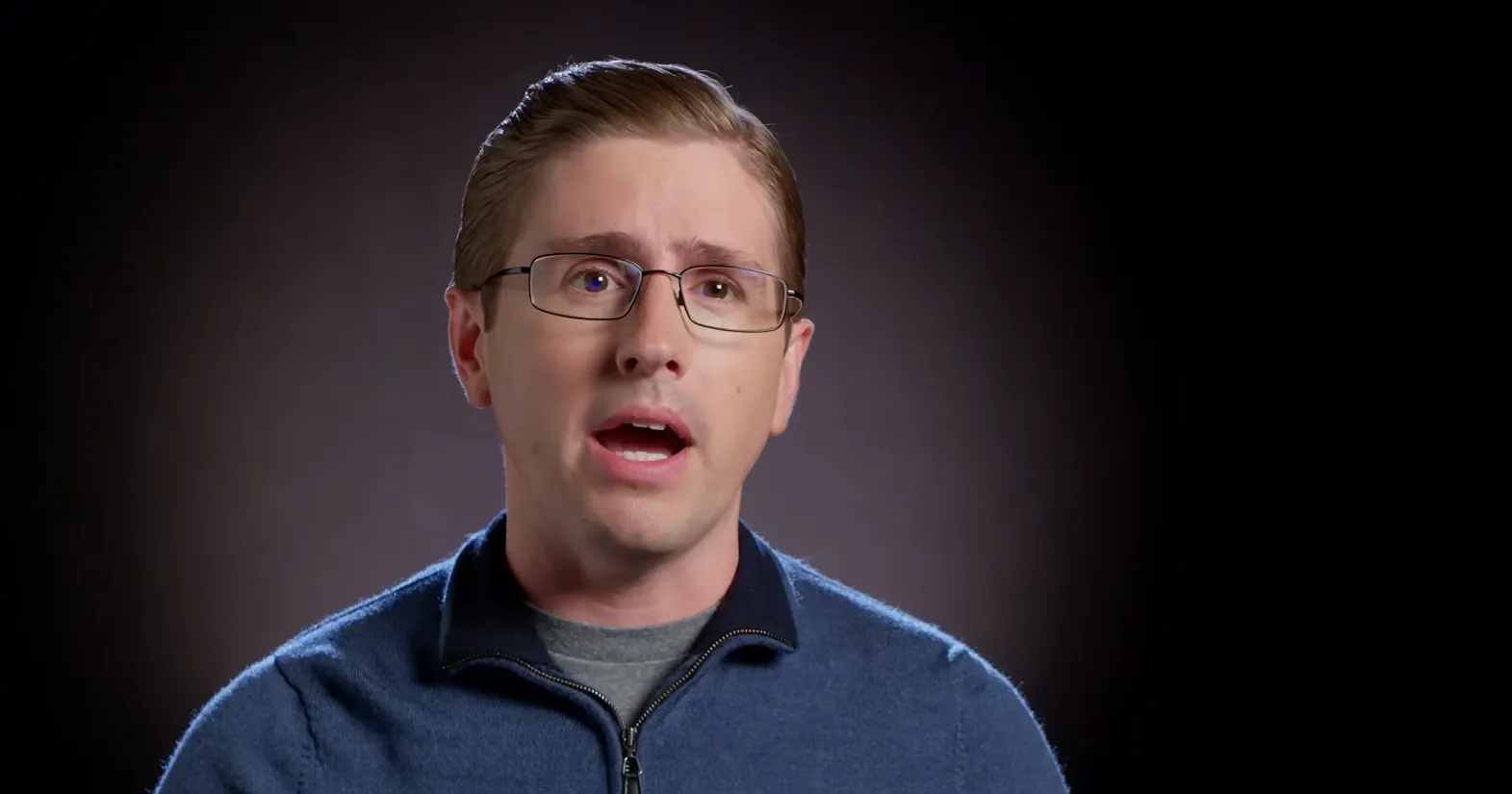 A headshot of a person in a blue sweater and eyeglasses on a black background.