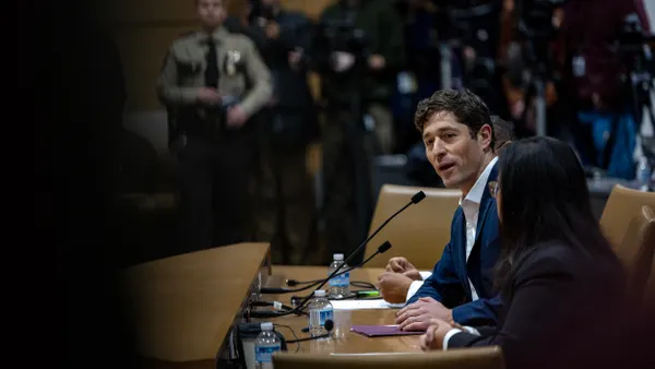 A person wearing a blue suit sits at a table speaking into a microphone while people sitting in seats surrounding look on.