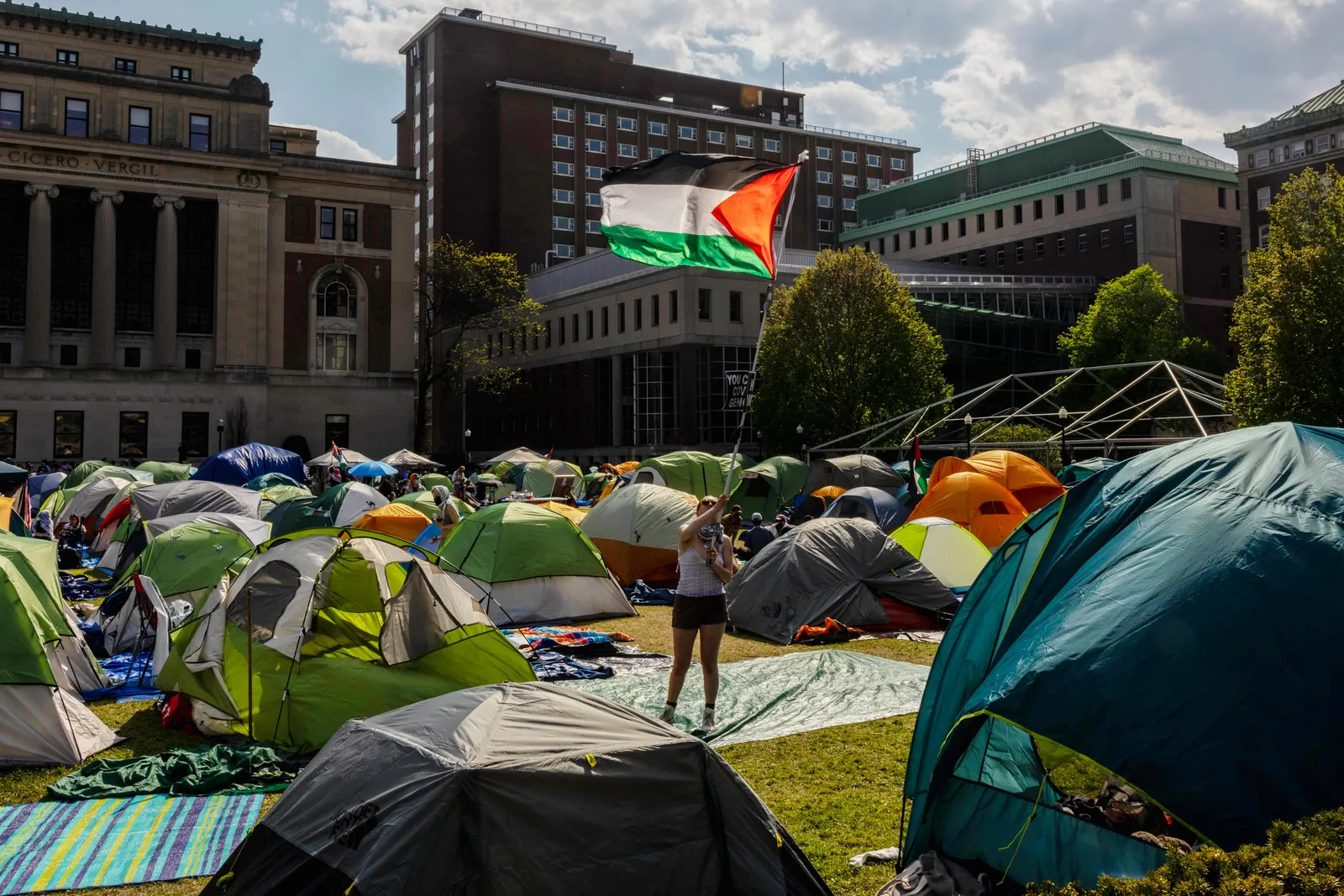 A person standing in the middle of multiple tents holds a flag, the background is a skyline of buildings.