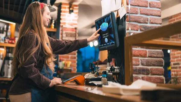 A person taps on a touch screen monitor that has a chatbot on it. The person is in a restaurant and wearing an apron.