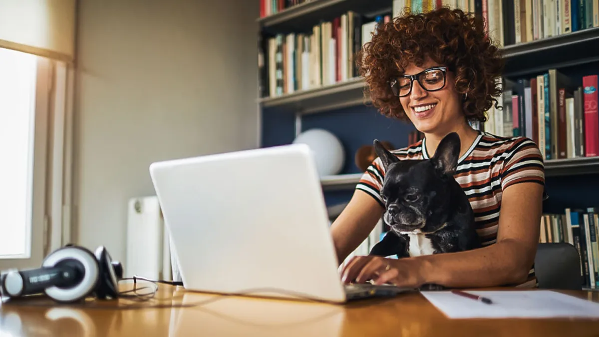 A person wearing glasses smiles at their laptop while a dog sits on their lap.