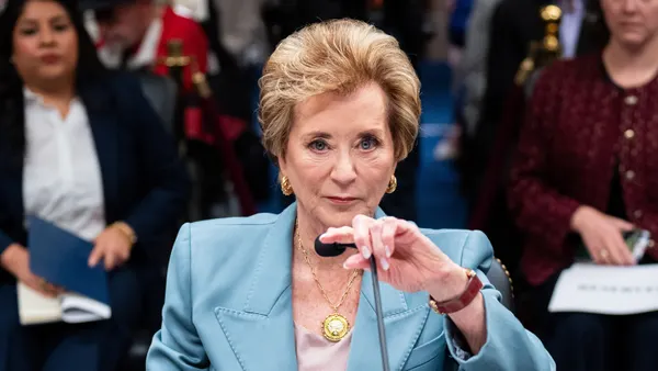 A person is sitting in a room with people sitting in rows behind them. The person is touch a microphone in front of them.