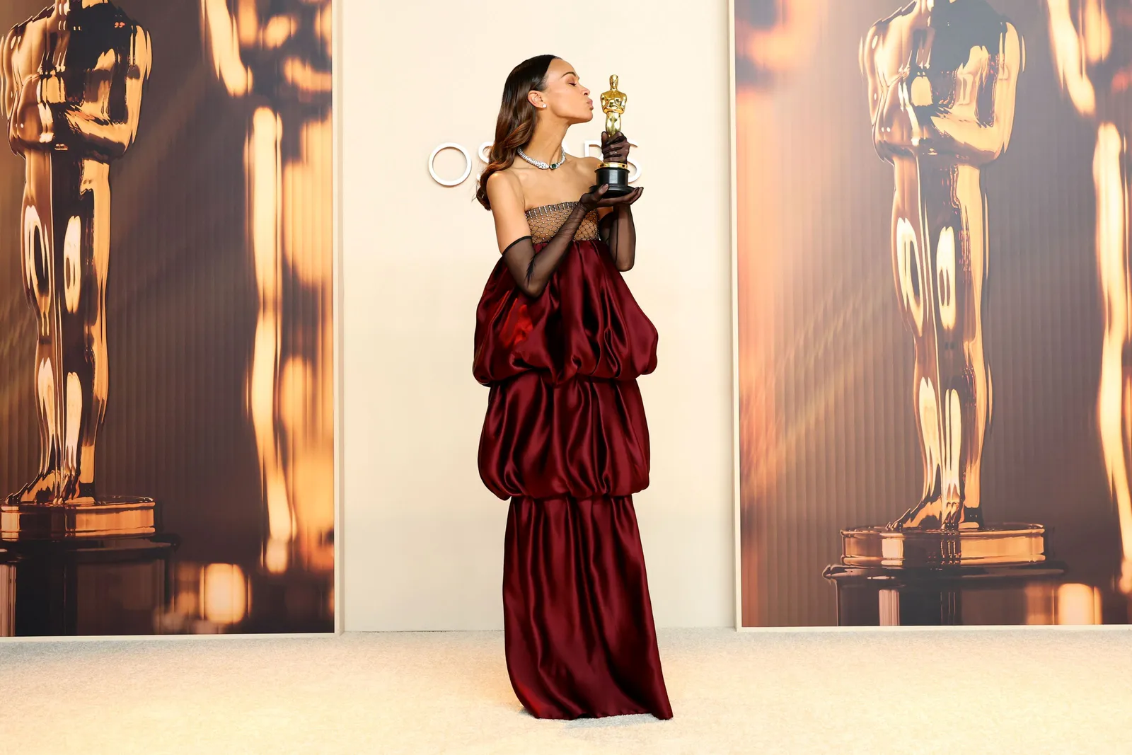 A woman in a burgundy bubble tiered dress with a sparkled mesh bodice and long sheer gloves stands on the red carpet and kisses the Oscar statue she holds.