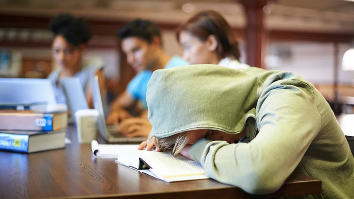 A student sitting at a desk is shown with their head down on a desk and their hood pulled over their head. Other students can be seen working in the background.