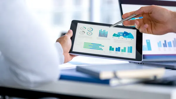 Two business men meeting and looking at financial data, charts and graphs on a digital tablet and a laptop computer. There are documents and paperwork on the table.