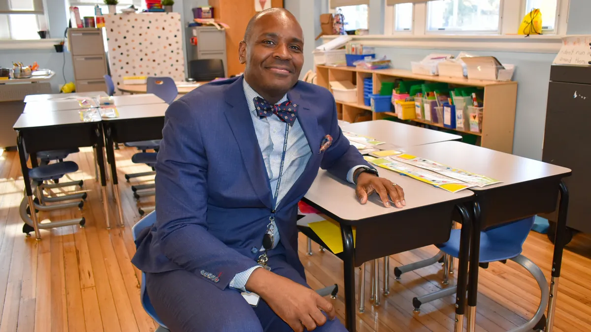 A person in a bow-tie and suit sits at a desk in an elementary school classroom and is looking at the camera. The classroom has windows, desks and a shelf of supplies.