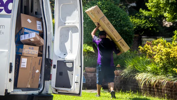 A person carries a large box in a neighborhood near a van with the back door open, showing more boxes.